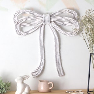 Pode incluir: Um grande laço branco tecido pendura-se numa parede branca. Um pequeno coelho de cerâmica branca, um jarro rosa e um vaso de metal preto com flores secas estão numa superfície de madeira sob o laço. Há também vegetação.