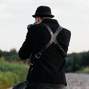 May include: A person kneels on a cobbled path, taking a photograph. They are dressed in black jacket, trousers, and shoes, with a black hat. A leather camera harness is visible. The background shows green grass and trees under a cloudy sky.