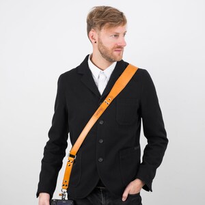 May include: A man wearing a black blazer and jeans is holding a black camera with a tan leather camera strap. The strap has the word "ORIGINAL" embossed on it.