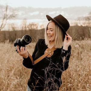 Op de afbeelding: Een vrouw in een zwart shirt, blauwe jeans en een zwarte fedora-hoed houdt een zwarte camera vast met een bruine leren cameraband. Ze staat in een veld met hoog gras.
