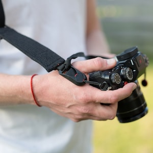 May include: A black camera strap with a red adjustable buckle. The strap is attached to a black camera with a lens.