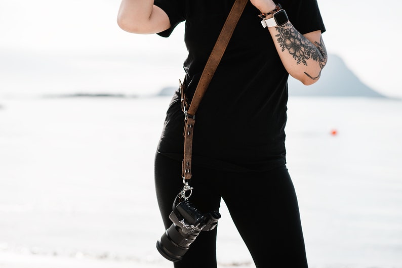 May include: A person wearing black clothing with a brown leather camera strap and a camera attached to the strap.