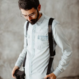 May include: A man wearing a light blue button-down shirt with black braces and the word "SHUZUMA" printed on one strap. He is holding two black cameras with straps.