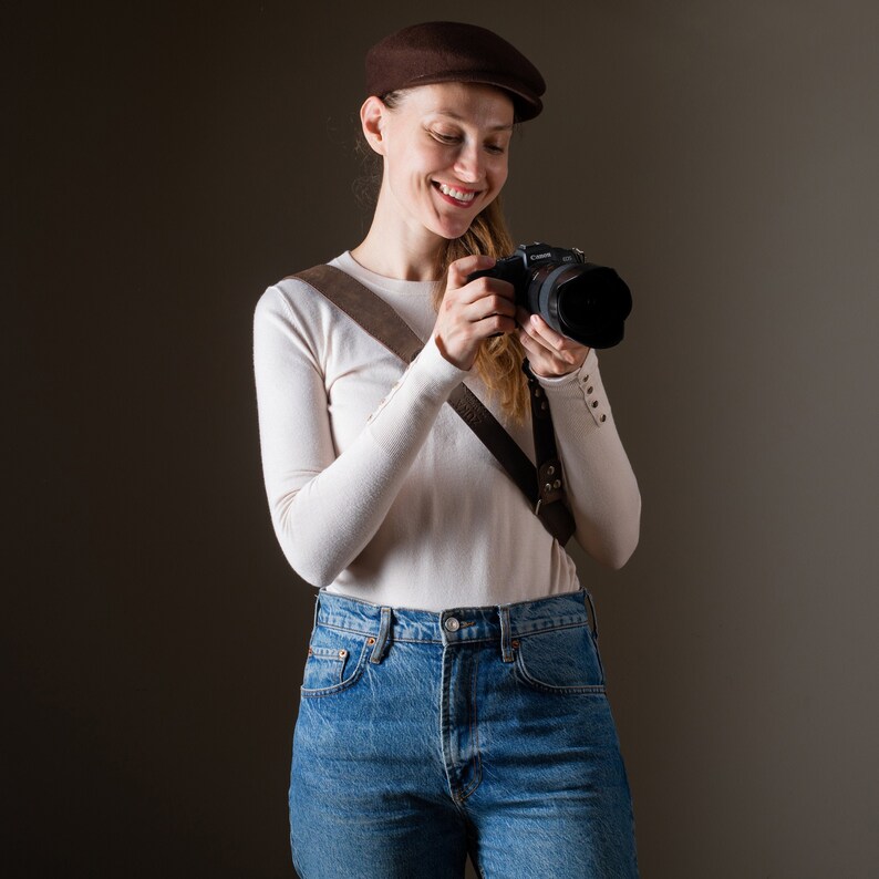 May include: A woman wearing a brown newsboy cap and a white sweater is holding a black camera with a brown leather strap. She is wearing blue jeans.