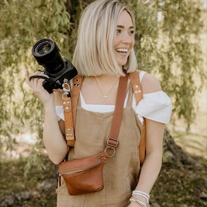 May include: A woman wearing a brown overall dress and a white shirt, holding a black camera with a brown leather strap. She is also wearing a brown leather fanny pack and a white bracelet.