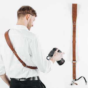 May include: A person wearing a white shirt and black pants is holding a camera with a brown leather camera strap. The strap is also shown hanging on a white wall.