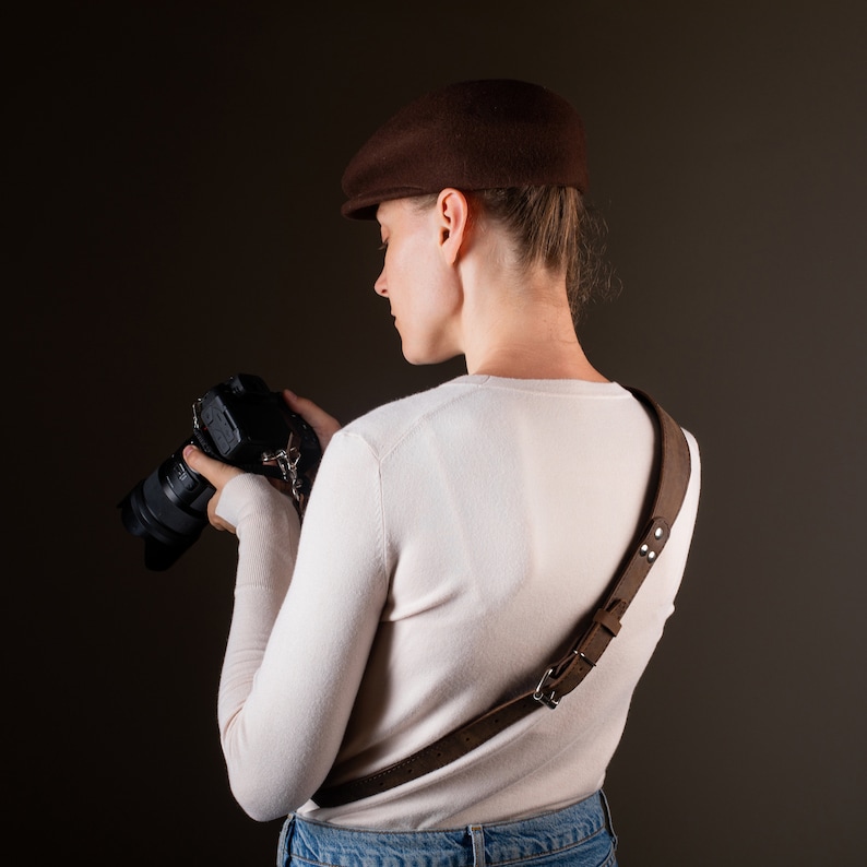 May include: A person wearing a brown newsboy cap and a white sweater is holding a black camera with a brown leather camera strap.