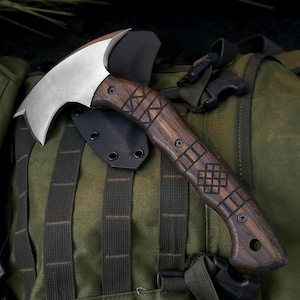 May include: A hand axe with a curved silver metal blade and a dark brown wooden handle featuring engraved geometric patterns. The axe includes a black kydex sheath with visible rivets, showcasing a sturdy construction and detailed wood grain.