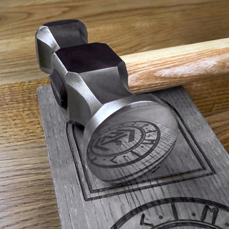 K&ouml;nnte beinhalten: Ein Stahlhammer mit Holzgriff liegt auf einem Holzblock mit einem eingravierten Design. Der Hammerkopf spiegelt das eingravierte Design wider. Der Hammer ist ein Werkzeug f&uuml;r Holz- oder Metallarbeiten.