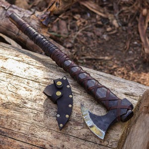 May include: A handcrafted axe with a dark wooden handle wrapped in brown leather. The axe head is made of dark metal with a polished blade. A matching leather sheath with brass rivets protects the blade. The axe is displayed on a wooden surface.