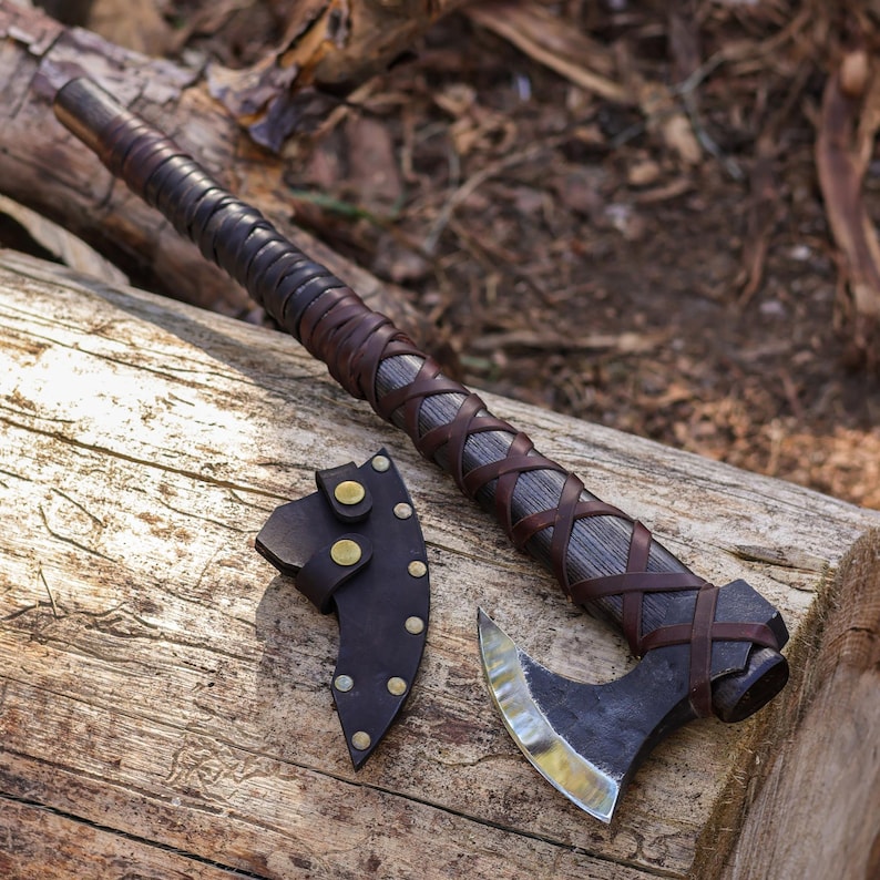 May include: A handcrafted axe with a dark wooden handle wrapped in brown leather. The axe head is made of dark metal with a polished blade. A matching leather sheath with brass rivets protects the blade. The axe is displayed on a wooden surface.