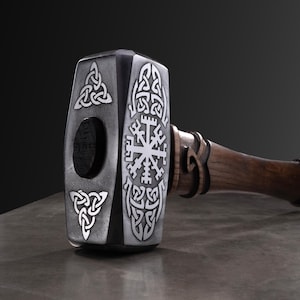 May include: A close-up of a hammer with a dark wooden handle and a metallic head. The head features intricate white Celtic knot designs and a circular hole. The hammer is a dark grey colour.