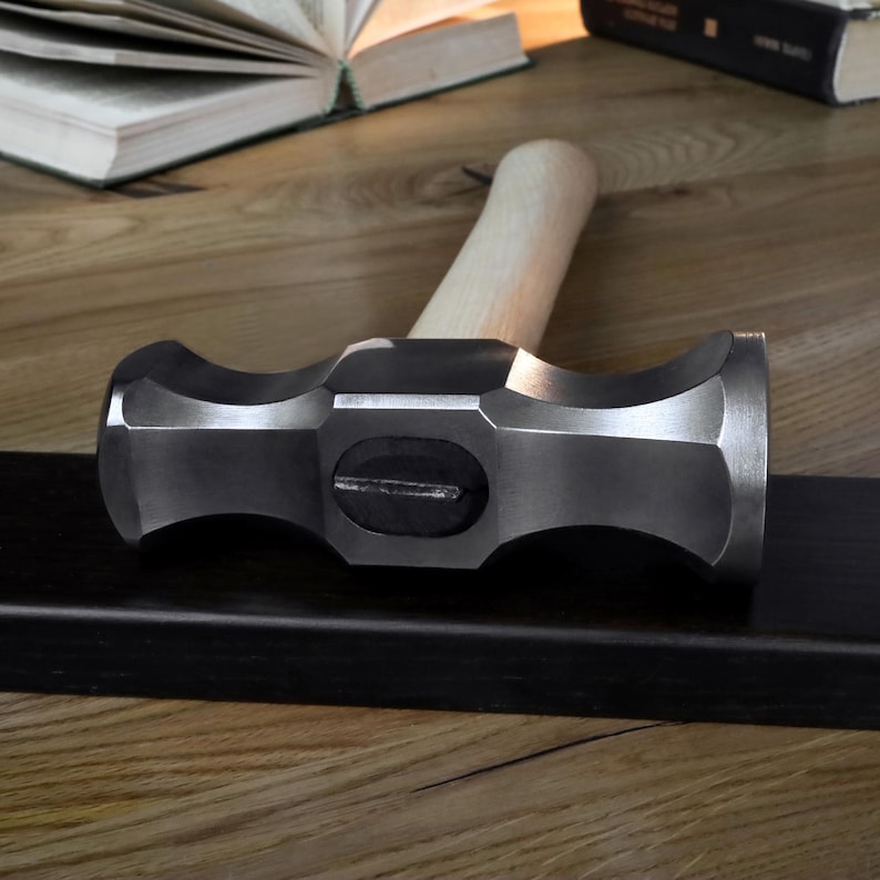 K&ouml;nnte beinhalten: Ein schwerer Vorschlaghammer aus Stahl mit einem hellen Holzgriff. Der Hammerkopf ist dunkelgrau und liegt auf einer schwarzen Oberfl&auml;che. Im Hintergrund befinden sich offene B&uuml;cher auf einem Holztisch.