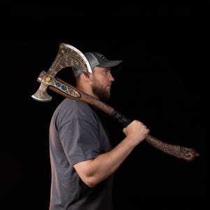 May include: A double-bitted battle axe featuring a dark, weathered metal head with intricate gold-colored filigree, three blue circular inlays, and a long, carved wooden handle with a textured grip and a decorative pommel shaped like a creature head.
