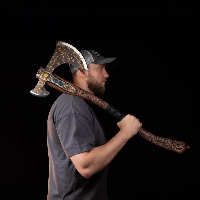 May include: A double-bitted battle axe featuring a dark, weathered metal head with intricate gold-colored filigree, three blue circular inlays, and a long, carved wooden handle with a textured grip and a decorative pommel shaped like a creature head.