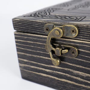 May include: A rectangular wooden box with a dark, distressed finish and visible wood grain. The lid features an engraved Celtic knot pattern. A brass-toned metal latch with a swing-arm mechanism is attached to the front of the box with small screws.