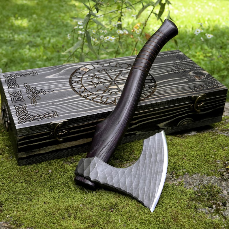 May include: A dark wood-handled axe with a textured, hammered metal blade resting on a rectangular wooden box. The box features a dark, charred finish with intricate laser-engraved Celtic knotwork and a circular Viking-style compass design.