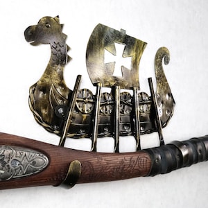 May include: A wall-mounted metal display rack shaped like a Viking longship with a gold and black finish, featuring a cutout cross on the sail. The rack includes four vertical hooks holding a wooden-handled weapon with ornate metal detailing.
