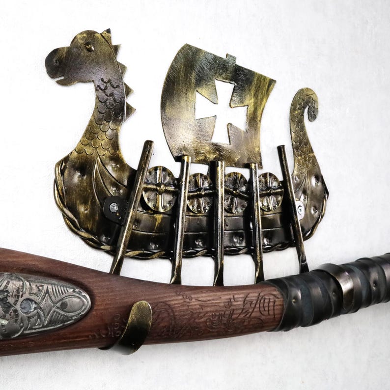 May include: A wall-mounted metal display rack shaped like a Viking longship with a gold and black finish, featuring a cutout cross on the sail. The rack includes four vertical hooks holding a wooden-handled weapon with ornate metal detailing.