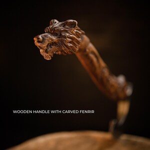 Pu&ograve; includere: Primo piano di un manico in legno con una testa di lupo scolpita. Il manico &egrave; di colore marrone scuro e la testa di lupo &egrave; finemente dettagliata. Il testo in basso recita "WOODEN HANDLE WITH CARVED FENRIR."