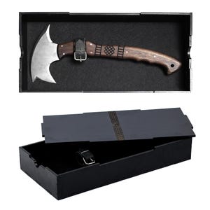 May include: A handcrafted axe with a curved, silver blade and a dark wooden handle. The handle features decorative carvings and is secured with a leather strap and buckle. The axe is presented in a black display box with a matching lid.
