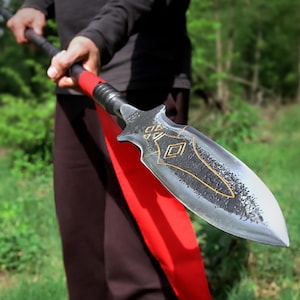 May include: A spear with a black shaft and a silver-coloured, pointed blade. The blade has a decorative gold-coloured design. A red ribbon is wrapped around the spear near the blade. The spear is held by a person.