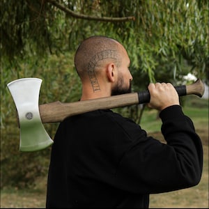 K&ouml;nnte beinhalten: Eine Doppelaxt mit polierter Stahlklinge und Holzgriff. Der Axtkopf hat ein kreisf&ouml;rmiges Emblem. Der Griff ist mit schwarzem Leder umwickelt. Die Axt wird &uuml;ber der Schulter getragen.