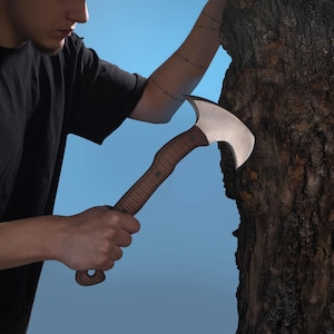 Könnte beinhalten: Eine silberne und braune Axt mit gebogener Klinge und Holzgriff. Die Axt wird in der Nähe eines Baumstamms gehalten. Der Griff hat einen strukturierten Griff und ist mit Nieten befestigt. Die Klinge ist gebogen und scharf.