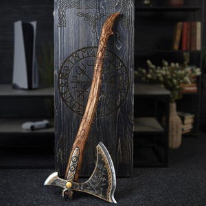 Puede incluir: Un hacha decorativa con mango de madera y hoja de metal. El mango está tallado intrincadamente con un acabado marrón claro, mientras que la hoja presenta grabados ornamentados. El hacha se exhibe contra un panel de madera oscura con símbolos tallados.