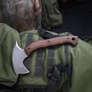 Op de afbeelding: Een handbijl met een gebogen, mat zilverkleurig metalen blad en een donkerbruin, getextureerd houten handvat. Het handvat heeft een reeks verticale groeven voor grip, is bevestigd met twee zwarte schroeven en bevat een rond gat aan het uiteinde.