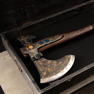 May include: A decorative battle axe with a dark metal head featuring gold-colored etched runic patterns and a sharp silver edge. The wooden handle has intricate carvings and a black leather strap with a metal buckle securing the axe head in place.