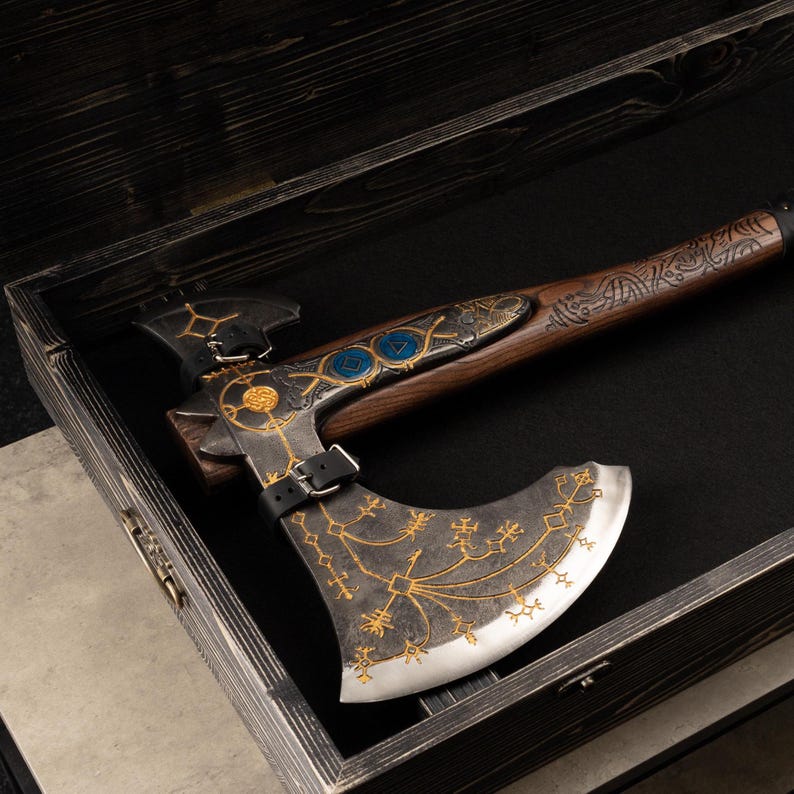 May include: A decorative battle axe with a dark metal head featuring gold-colored etched runic patterns and a sharp silver edge. The wooden handle has intricate carvings and a black leather strap with a metal buckle securing the axe head in place.