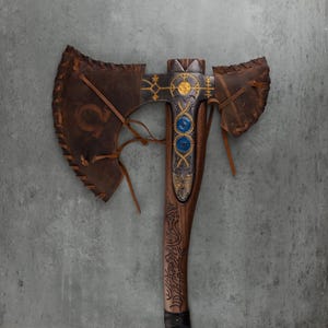 May include: A double-bitted axe featuring a dark brown wooden handle with intricate carvings, a metal head with gold-colored geometric engravings, two circular blue inlays, and brown leather covers laced onto the blades with leather cords.