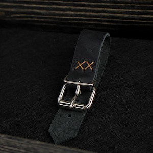 May include: A black leather strap featuring a silver-toned metal buckle and a pointed end. The strap is secured with two distinct cross-stitch patterns in a light brown thread, positioned above the buckle hardware on the main leather loop.