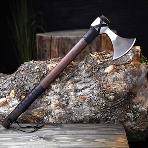 May include: A handcrafted axe with a polished steel blade and a dark wooden handle. The handle is wrapped with black leather for grip. The axe is displayed on a textured, weathered log, showcasing its rustic design.