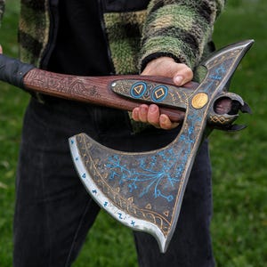 Puede incluir: Un hacha decorativa detallada con un mango de madera y una hoja de metal. La hoja presenta intrincados diseños azules y dorados, con un acabado gris oscuro. El mango está envuelto en cinta negra y tiene detalles tallados.