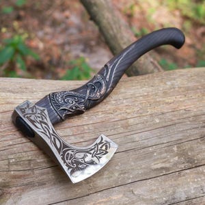 May include: A handcrafted axe with a dark wooden handle and a silver-colored blade. The handle features intricate dragon and knotwork carvings. The axe is displayed on a weathered wooden surface, showcasing its detailed craftsmanship.