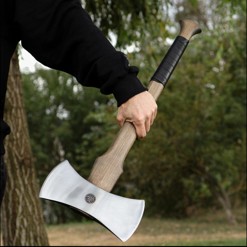 K&ouml;nnte beinhalten: Eine Doppelaxt mit Holzgriff und silberfarbener Klinge. Der Griff ist oben mit schwarzem Leder umwickelt. Ein kreisf&ouml;rmiges Emblem ist auf der Klinge sichtbar. Die Axt wird in der Hand einer Person gehalten.