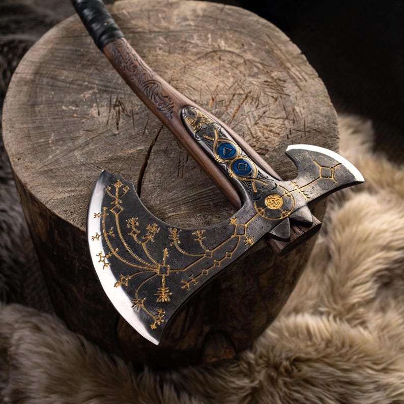 May include: A double-bitted axe with a dark metal head featuring intricate gold-colored etched patterns and two blue circular inlays. The handle is made of brown wood with carved details and a black wrapped grip near the top of the shaft.