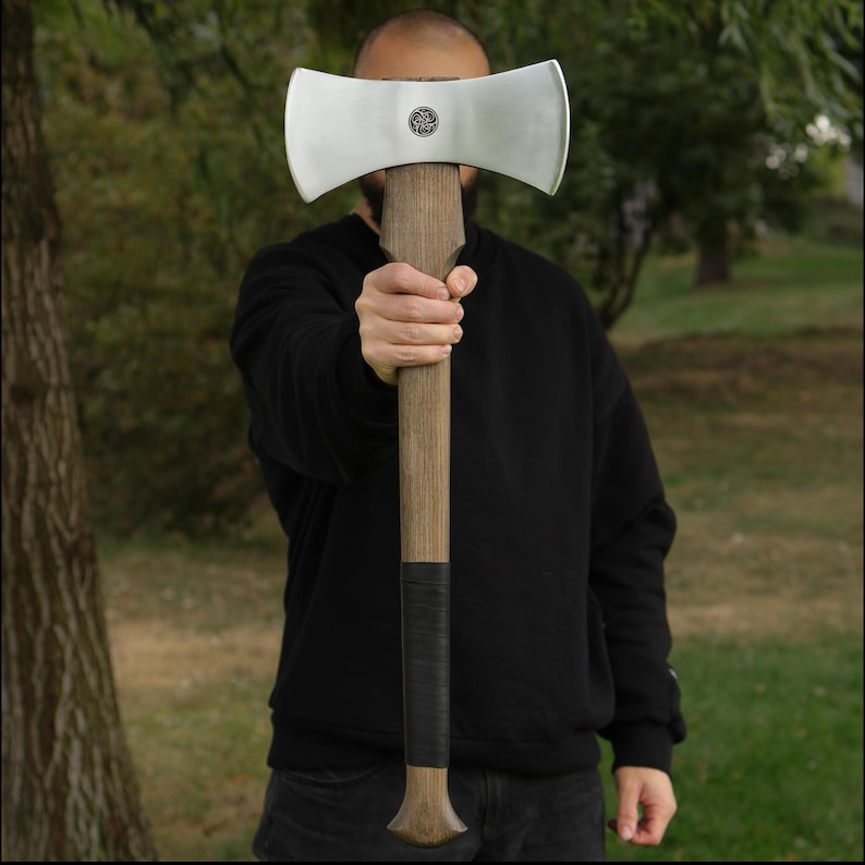 K&ouml;nnte beinhalten: Eine Doppelaxt mit polierter Stahlklinge und Holzgriff. Der Axtkopf hat ein dekoratives Emblem. Der Griff ist mit schwarzem Material umwickelt, um den Halt zu verbessern. Die Axt wird vor einer Person gehalten.