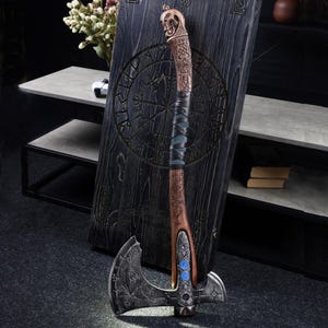 May include: A decorative axe with a detailed, engraved blade and a wooden handle. The handle is wrapped with dark blue leather and features intricate carvings. The axe is displayed against a dark wooden panel with etched designs, suggesting a Viking or fantasy theme.