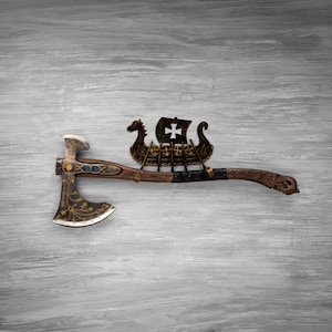 May include: A decorative battle axe featuring a dark metal head with gold filigree patterns and blue inlays. The wooden handle is adorned with a metal Viking longship ornament, complete with a sail displaying a cross, and intricate carvings.