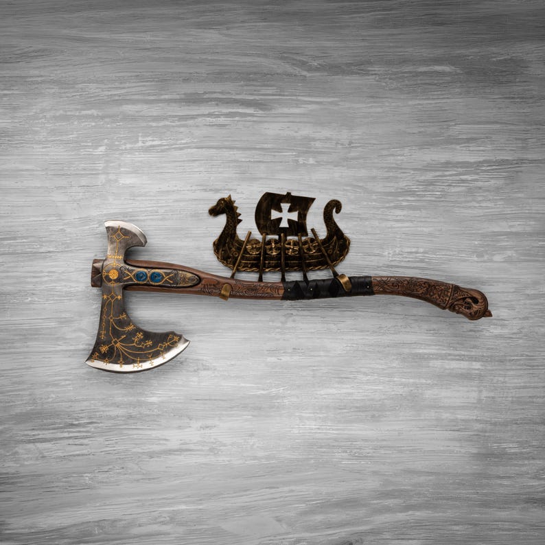 May include: A decorative battle axe featuring a dark metal head with gold filigree patterns and blue inlays. The wooden handle is adorned with a metal Viking longship ornament, complete with a sail displaying a cross, and intricate carvings.