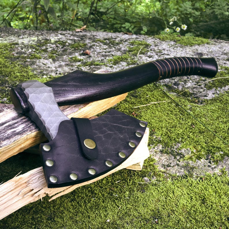 May include: A hand axe with a textured, grey metal head and a curved, dark black wooden handle. The axe blade is partially covered by a black leather sheath secured with brass-colored metal rivets and a central snap button closure.