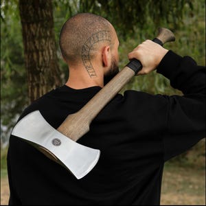 May include: A double-bit axe with a polished steel blade and a wooden handle. The axe head features a circular emblem. The handle is a light brown color with a black grip near the head. The axe is held over a person's shoulder.