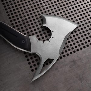 Puede incluir: Un hacha plateada con un mango negro. La cabeza del hacha tiene un dise&ntilde;o &uacute;nico con un recorte circular y una hoja puntiaguda. El mango es negro con un agarre texturizado. El hacha est&aacute; sobre una superficie met&aacute;lica gris perforada.