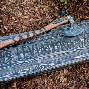 Puede incluir: Un hacha artesanal con mango de madera y hoja azul y dorada, apoyada sobre una caja de madera. La caja está grabada con las palabras "LEVIATHAN AXE" y un símbolo náutico. El mango del hacha tiene detalles decorativos negros.
