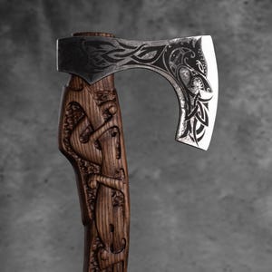 May include: Axe with a dark wooden handle featuring carved relief artwork. The axe head is silver with etched designs. The blade has a curved shape. The overall design suggests a Viking or Norse theme.
