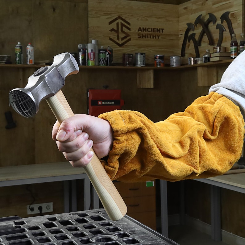 K&ouml;nnte beinhalten: Ein Metallhammer mit Holzgriff wird in einer Hand gehalten, die einen gelben Schutz&auml;rmel tr&auml;gt. Der Hammer hat einen abgerundeten Kopf mit strukturierter Oberfl&auml;che. Im Hintergrund sind die Worte "ANCIENT SMITHY" zu sehen.