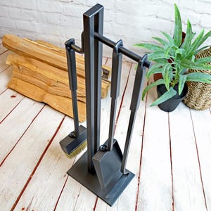 Puede incluir: Un juego de herramientas para chimenea de metal negro con un soporte. El juego incluye un cepillo, una pala, un atizador y un elevador de troncos. Las herramientas se exhiben sobre una base cuadrada. Leña y una planta en maceta en el fondo.